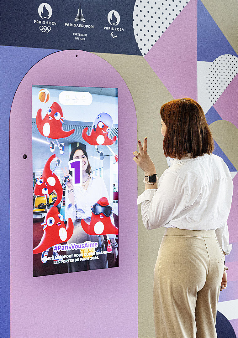 A passenger poses in front of an augmented reality screen featuring Paris 2024 visuals.