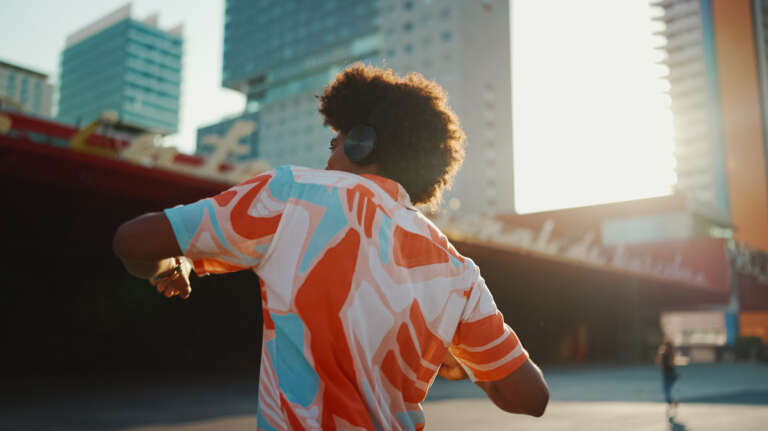 A young man wearing a colorful shirt dances on the street and has headphones on his head.